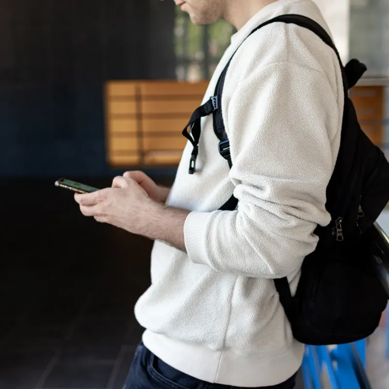 Person in a white jumper and backpack checking a phone by a railing, head out of frame